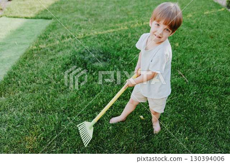 Playful boy working in the garden, enjoying outdoor activities with a bright smile, wearing casual clothes and standing on lush green grass in the sunlight. 130394006