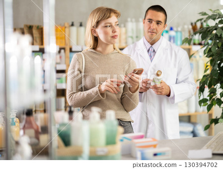 Girl client scrolls phone and shopping in pharmacy, male pharmacist in background. Girl client scrolls phone and shopping in pharmacy, male pharmacist in background. 130394702