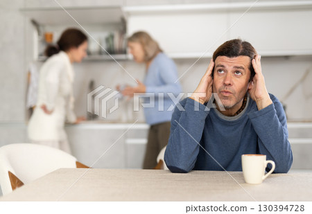 Frustrated man sitting in kitchen during quarrel between his wife and elderly mother. 130394728
