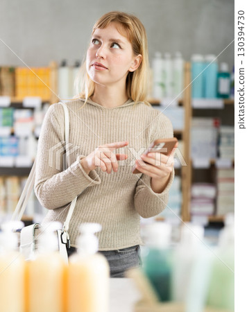 Female shopper searches for necessary goods on the shelves of a supermarket, checking the list on smartphone 130394730