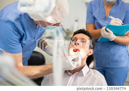 During examination of male patients teeth, nurse listens to senior male doctors dentist instructions 130394774