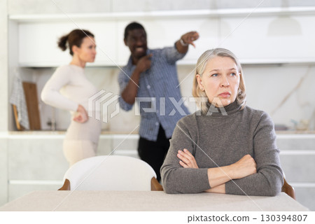 Old woman sitting at the kitchen table with her back to couple quarreling together 130394807