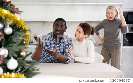 Middle-aged couple sitting at table and mature woman scolding them in the kitchen with Christmas-tree 130394977