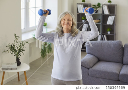 Smiling sporty older woman lifting dumbbells overhead training alone at home demonstrating lifestyle 130396669