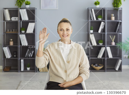 Woman smiling and waving at camera from home or office online, background of tall shelves with books Woman smiling and waving at camera from home or office online, background of tall shelves with books 130396670