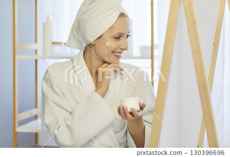 Young woman looking at mirror applying moisturizing cream on face doing morning beauty procedure Young woman looking at mirror applying moisturizing cream on face doing morning beauty procedure 130396696