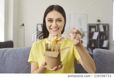 Happy young woman sitting on couch, eating noodles with chopsticks and enjoying tasty takeaway food. Happy young woman sitting on couch, eating noodles with chopsticks and enjoying tasty takeaway food. 130396713