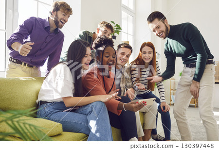 Diverse group of happy friends sitting on sofa using mobile phone at home together. 130397448