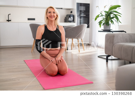 Blonde woman in sportswear sitting cross legged on yoga mat in modern living room smiling after yoga session, feeling calm and satisfied Blonde woman in sportswear sitting cross legged on yoga mat in modern living room smiling after yoga session, feeling calm and satisfied 130398268