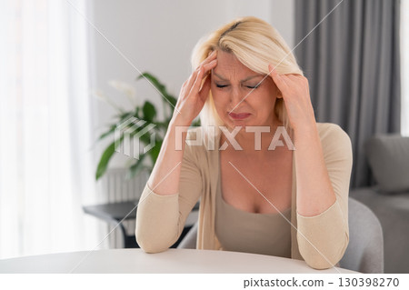 Blonde middle aged woman sitting alone at kitchen table, covering face with hands, crying with despair and sadness, emotional breakdown at home 130398270
