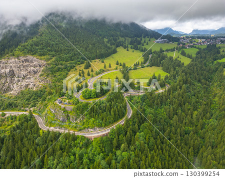 This aerial view showcases winding asphalt roads through the lush green hills of the Alps, with dramatic cloud cover enhancing the serene beauty of a road trip adventure. 130399254