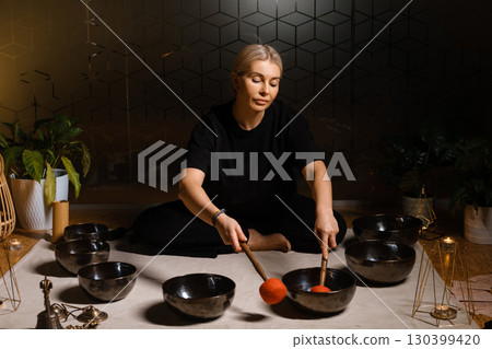Woman practicing sound healing with singing bowls in a tranquil indoor space decorated with plants and soft lighting during a peaceful evening session 130399420