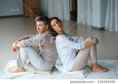 Couple engaging in mindfulness meditation practice while seated back to back in a serene indoor space during the late afternoon light 130399456