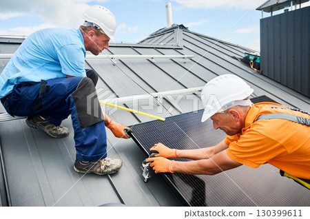Workers building photovoltaic solar panel system on rooftop of house. Men technicians in helmets and gloves installing solar module with help of hex key outdoors. Renewable energy generation concept. 130399611