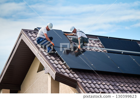 Workers building solar panel system on rooftop of house. Two men installers in helmets installing photovoltaic solar module outdoors. Alternative, green and renewable energy generation concept. 130399623