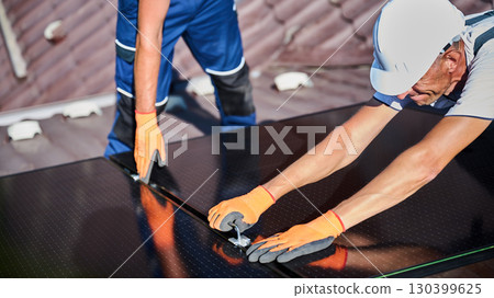 Workers building photovoltaic solar panel system on rooftop of house. Close up of men technicians in helmets and gloves installing solar module with help of hex key outdoors. Alternative energy. Workers building photovoltaic solar panel system on rooftop of house. Close up of men technicians in helmets and gloves installing solar module with help of hex key outdoors. Alternative energy. 130399625