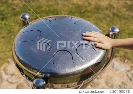 Hand rests on the metallic surface of a musical instrument while a child enjoys creating sounds in a vibrant park under clear blue skies in the afternoon Hand rests on the metallic surface of a musical instrument while a child enjoys creating sounds in a vibrant park under clear blue skies in the afternoon 130400078