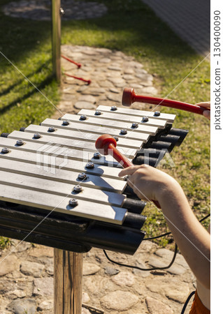Children are having fun creating melodies on a xylophone with bright mallets in a lively outdoor park. The sun illuminates their joyful expressions and the surrounding green grass Children are having fun creating melodies on a xylophone with bright mallets in a lively outdoor park. The sun illuminates their joyful expressions and the surrounding green grass 130400090