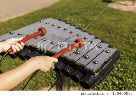 Two hands grasp red mallets, striking metal bars of a musical instrument in a park. Sunlight casts a warm glow, highlighting the playful atmosphere for children and families Two hands grasp red mallets, striking metal bars of a musical instrument in a park. Sunlight casts a warm glow, highlighting the playful atmosphere for children and families 130400093