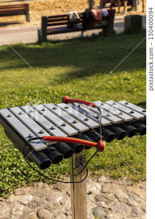 A bright xylophone rests on a wooden post in a lively park, surrounded by green grass and wooden benches. People enjoy their time nearby on a sunny afternoon A bright xylophone rests on a wooden post in a lively park, surrounded by green grass and wooden benches. People enjoy their time nearby on a sunny afternoon 130400094