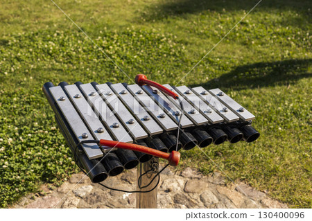 Colorful musical instrument featuring metal bars and tubes stands in a green park. Red mallets rest on the bars, inviting playful music making in a sunny environment Colorful musical instrument featuring metal bars and tubes stands in a green park. Red mallets rest on the bars, inviting playful music making in a sunny environment 130400096