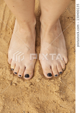 Feet with dark nail polish stand on warm sand at the beach, basking in the sunlight. Soft grains surround them, evoking feelings of tranquility and connection to nature Feet with dark nail polish stand on warm sand at the beach, basking in the sunlight. Soft grains surround them, evoking feelings of tranquility and connection to nature 130400135