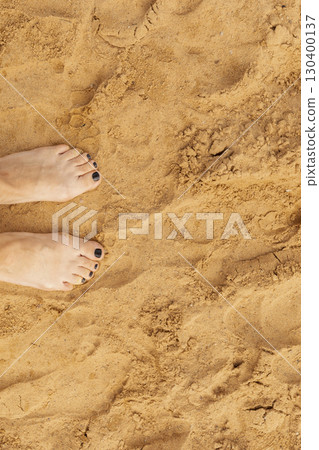 Two bare feet with dark nail polish stand on golden sand, basking in sunlight at a beach, surrounded by the warmth and tranquility of summer Two bare feet with dark nail polish stand on golden sand, basking in sunlight at a beach, surrounded by the warmth and tranquility of summer 130400137