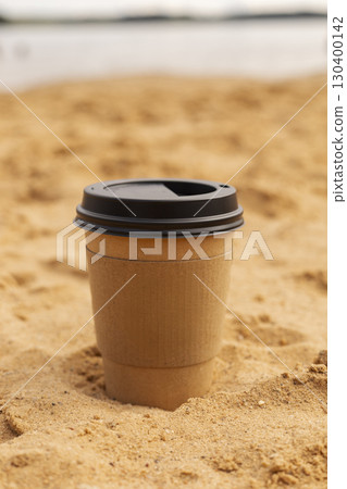 A single paper coffee cup sits partially buried in golden sand, with gentle waves visible in the background under a clear sky. It captures a serene moment by the beach 130400142