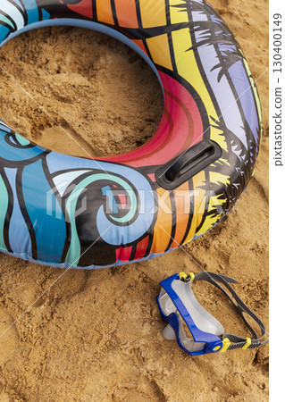 An inflatable tube with vibrant patterns rests on warm sand beside a pair of swimming goggles, inviting beachgoers to enjoy a sunny day filled with water activities and relaxation An inflatable tube with vibrant patterns rests on warm sand beside a pair of swimming goggles, inviting beachgoers to enjoy a sunny day filled with water activities and relaxation 130400149