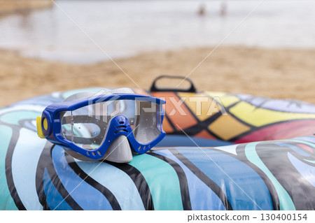 Bright blue swimming goggles sit on a colorful beach float near the water, with a sandy area and other beachgoers in the background, enjoying a sunny afternoon 130400154