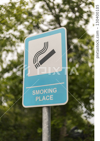 The sign shows a clear symbol and text indicating a smoking place, positioned among lush trees on a cloudy day, guiding smokers to the designated area 130400185