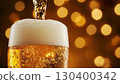 Close-up of cold lager pouring into a frosted glass with thick foam against a festive bokeh background. Festive setting with lights in the background, no people. Festive food concept for Oktoberfest. 130400342