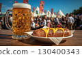 Close-up of refreshing cold foamy light beer and Bavarian sausage topped with mustard on a wooden table at an Oktoberfest celebration. Blurred people having fun, tents and flags in the background. 130400343