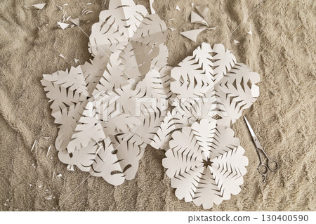 Snowflakes made from white paper lie on a beige fabric surface, surrounded by small paper scraps. A pair of scissors rests nearby, indicating recent crafting activities 130400590