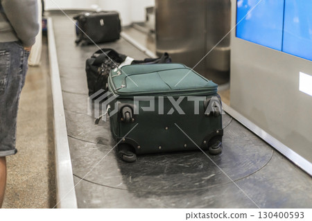 A green suitcase rests on the baggage carousel at an airport during a bustling evening. Nearby travelers are waiting for their luggage, creating a busy atmosphere as the journey continues 130400593