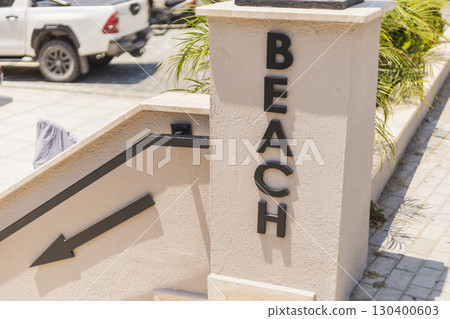 Clear beach access sign guides visitors down the steps toward the sandy shore. Palm plants line the path, creating a relaxed seaside ambiance as vehicles park nearby 130400603