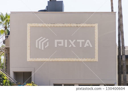 A beige building features an ornate wall frame design surrounding a blank area. Bright sunlight highlights the decorative elements, and palm trees add a tropical touch to the environment 130400604
