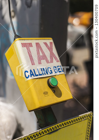 A bright yellow calling bell marked 'TAX' and 'CALLING BELL' hangs in a bustling urban area, indicating a call for taxi service. The vibrant color catches attention against the backdrop of city life A bright yellow calling bell marked 'TAX' and 'CALLING BELL' hangs in a bustling urban area, indicating a call for taxi service. The vibrant color catches attention against the backdrop of city life 130400709