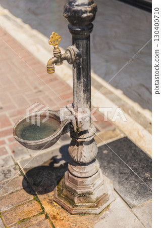 The antique water fountain stands proudly on a brick walkway, with crystal-clear water collecting in its basin. Sunlight illuminates the surrounding foliage, creating a peaceful atmosphere 130400710