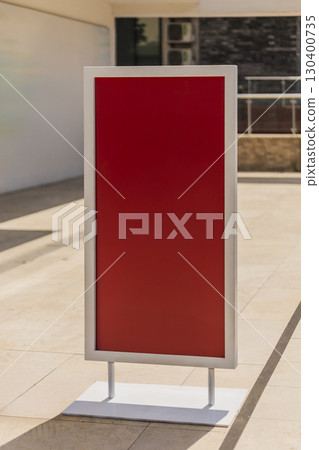 A blank red sign stands upright on a metal base in a well-lit outdoor area. The sleek design contrasts with the light-colored stone flooring and nearby building features A blank red sign stands upright on a metal base in a well-lit outdoor area. The sleek design contrasts with the light-colored stone flooring and nearby building features 130400735