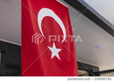 A vibrant red Turkish flag with a white star and crescent is hanging outside a modern building. The setting is lively, with bright sunlight shining down on the flag 130400748