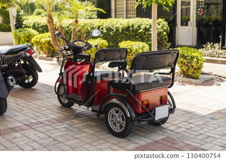 Brightly colored electric tricycle is parked under palm trees in a vibrant urban setting. The well-maintained area has greenery, suggesting a warm day ideal for outdoor activities Brightly colored electric tricycle is parked under palm trees in a vibrant urban setting. The well-maintained area has greenery, suggesting a warm day ideal for outdoor activities 130400754