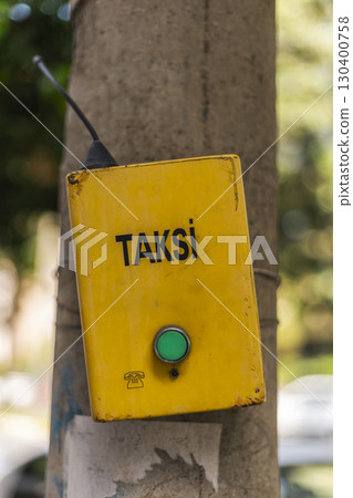 A bright yellow taxi call box is affixed to a tree trunk in a busy city area. The clear green button invites commuters to request a taxi service as cars pass by A bright yellow taxi call box is affixed to a tree trunk in a busy city area. The clear green button invites commuters to request a taxi service as cars pass by 130400758