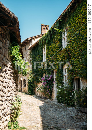 Ivy-covered stone house with blooming flowers along a cobblestone street under bright summer light. Rural French aesthetic, countryside charm, romantic facades, poetic living Ivy-covered stone house with blooming flowers along a cobblestone street under bright summer light. Rural French aesthetic, countryside charm, romantic facades, poetic living 130400947