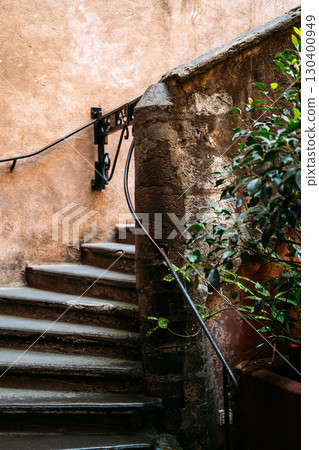 Worn stone staircase with iron railing curves up beside textured peach-colored wall and potted greenery. Urban decay, historic preservation, architectural detail, Mediterranean ambiance 130400949