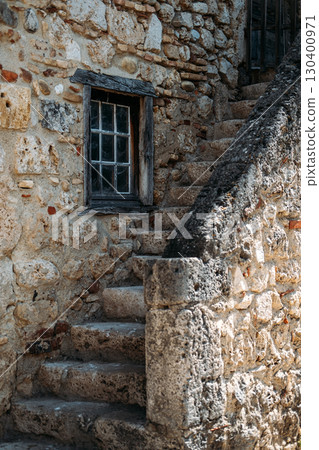 Aged stone staircase beside a rustic window framed by worn wooden beams on an old stone wall. Quiet living, countryside serenity, escapist domesticity, contemplative design Aged stone staircase beside a rustic window framed by worn wooden beams on an old stone wall. Quiet living, countryside serenity, escapist domesticity, contemplative design 130400971
