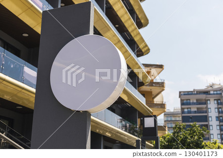 Large blank circular sign stands on a sturdy pole, set against a backdrop of contemporary buildings with colorful facades. The scene captures the essence of urban development. Round signboard mockup. Large blank circular sign stands on a sturdy pole, set against a backdrop of contemporary buildings with colorful facades. The scene captures the essence of urban development. Round signboard mockup. 130401173