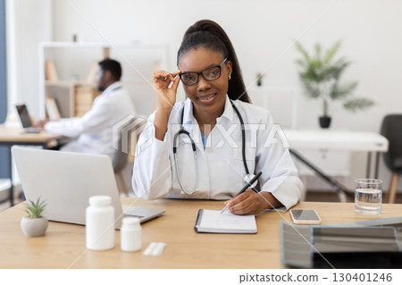 Female doctor working in office writing prescription on notepad using laptop for research 130401246