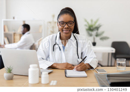 Young adult African female doctor in professional office writing prescription using laptop and notepad. Medical professional wearing white coat with stethoscope at work, healthcare 130401249