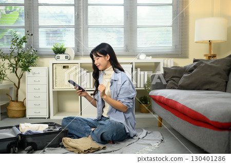 Young woman sitting on the floor, checking smartphone while packing clothes into a suitcase for travel Young woman sitting on the floor, checking smartphone while packing clothes into a suitcase for travel 130401286
